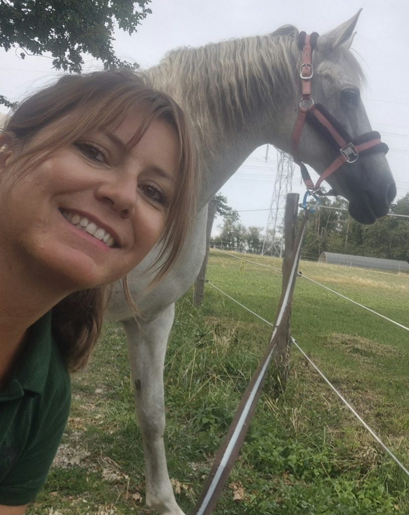 Lydie Autrand, ostéopathe animalier, souriante auprès d'un cheval gris dans son pré
